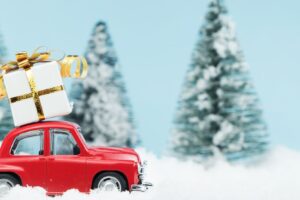 A red toy car with a gift box on the roof with snow on the ground and evergreen trees in the background