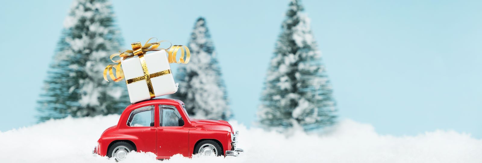A red toy car with a gift box on the roof with snow on the ground and evergreen trees in the background